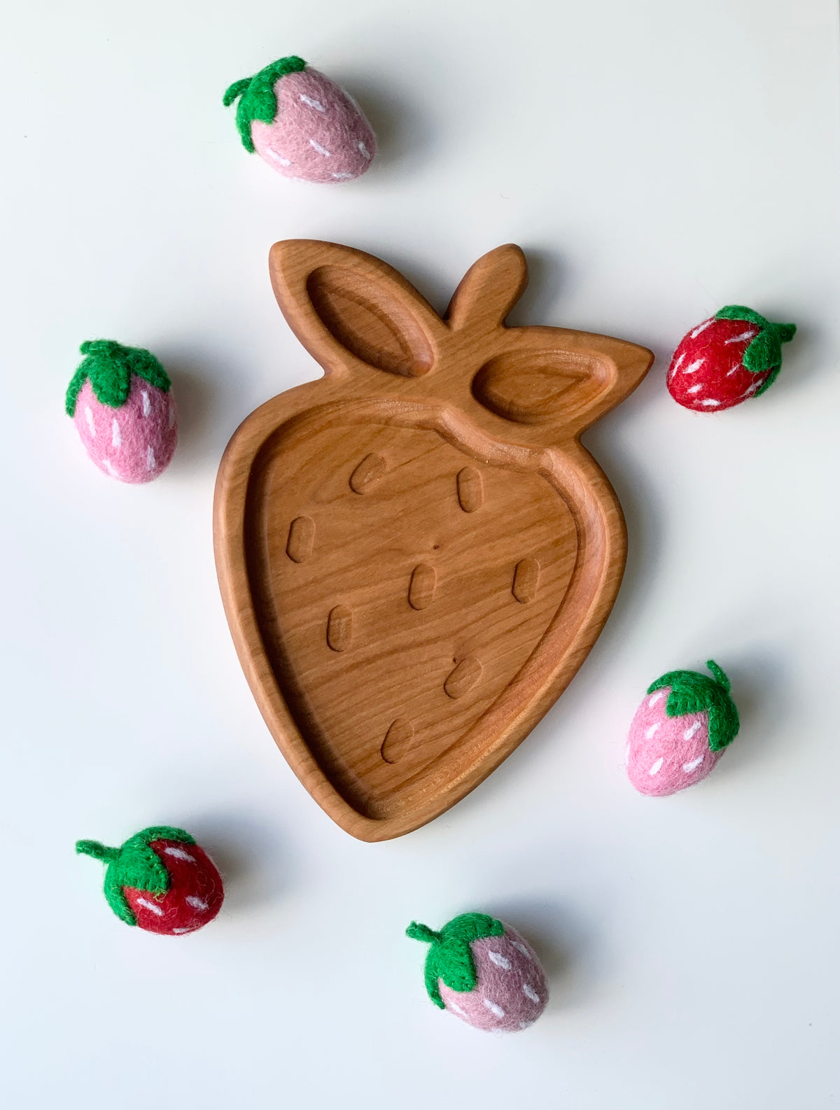 Cherry wood strawberry-shaped plate with carved seed details surrounded by small felt strawberries on white background