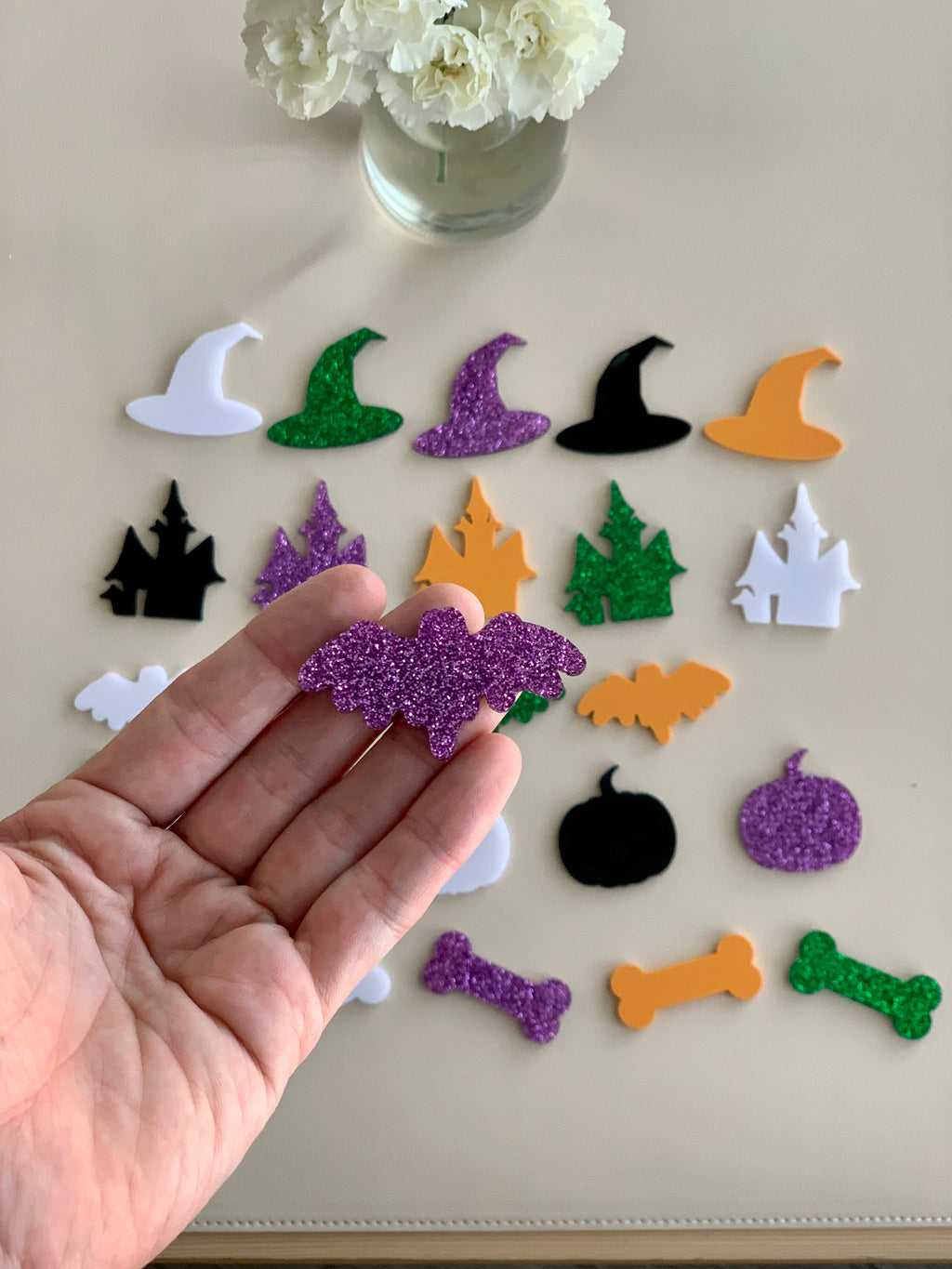 Hand holding purple glitter bat acrylic shape with various colored Halloween acrylic shapes spread on beige surface and white flowers in vase