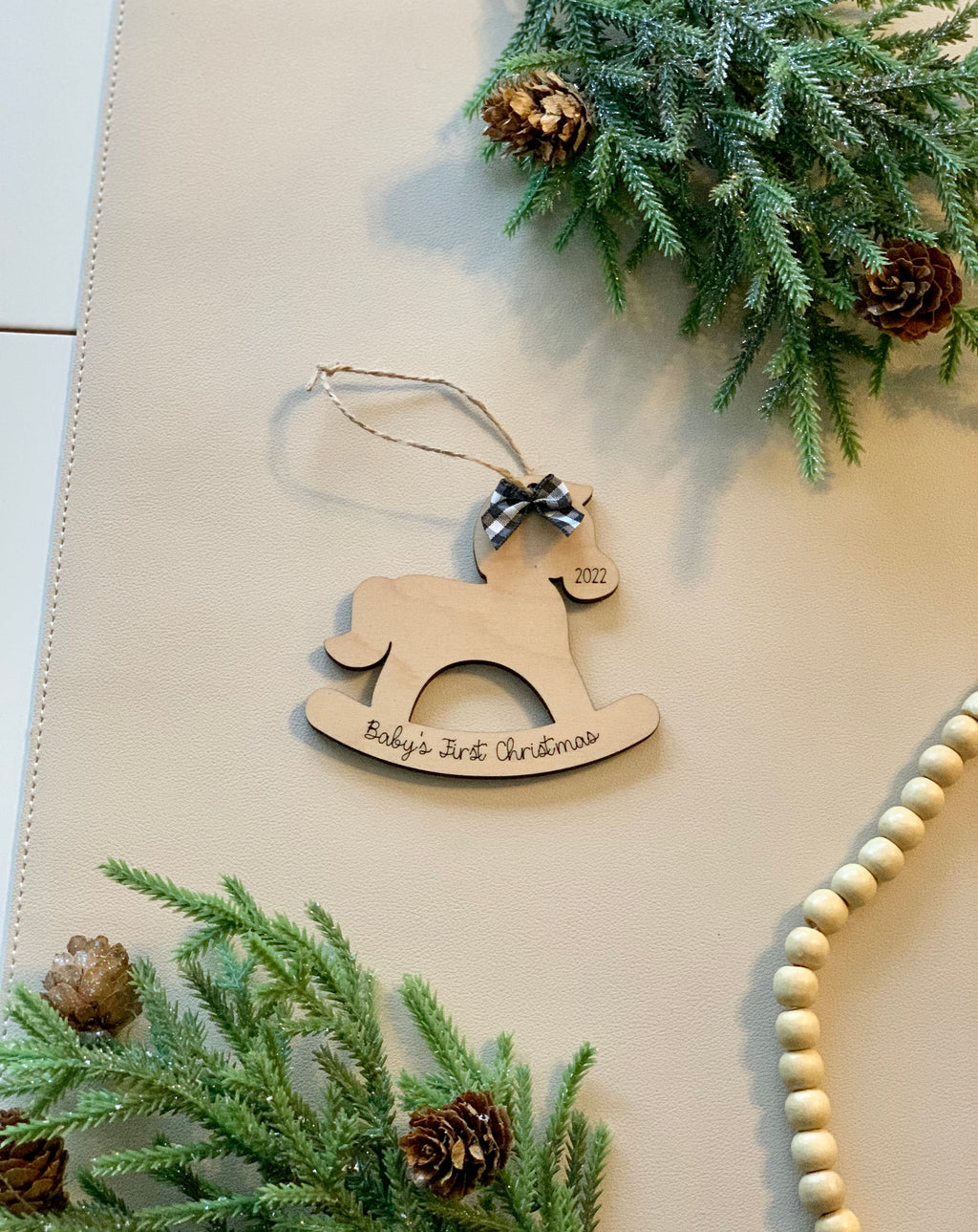 Wooden rocking horse Christmas ornament with “Baby’s First Christmas” inscription and a black checkered bow on beige surface with pine accents