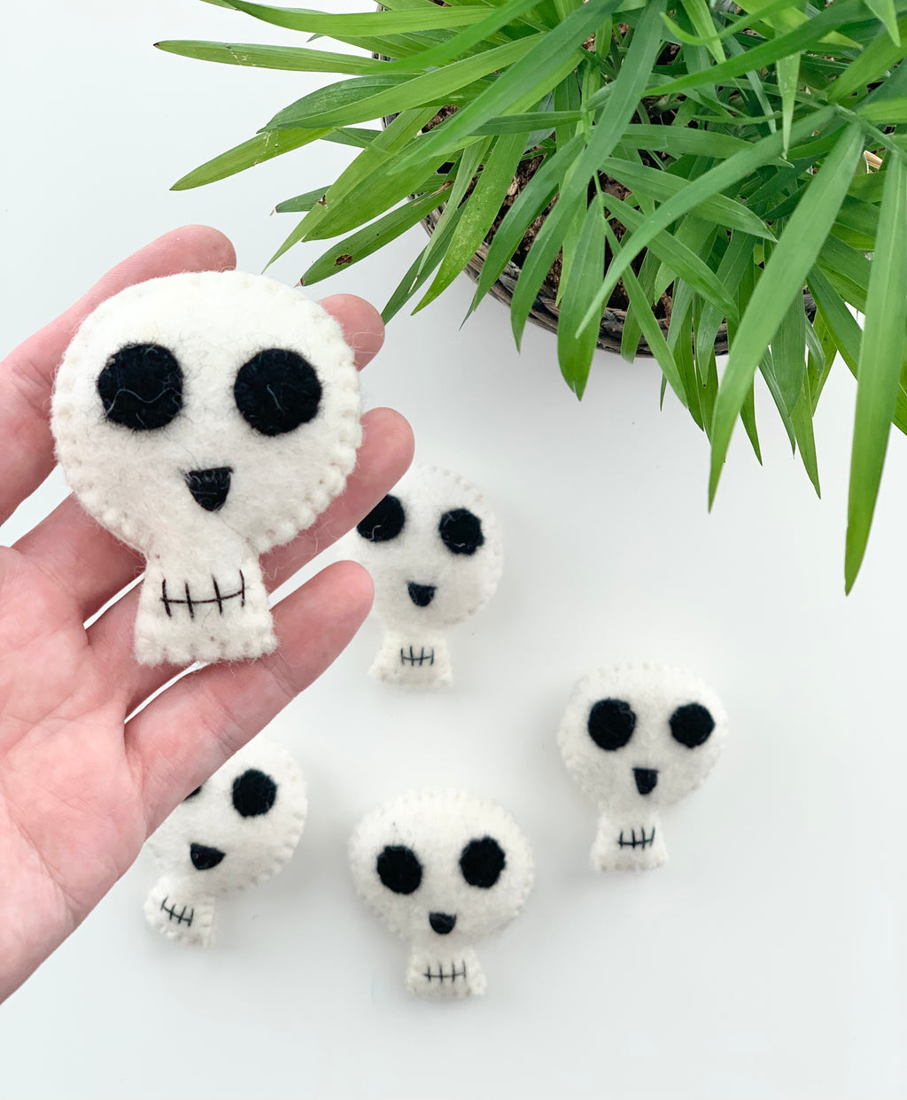 Handmade small white felt Halloween skull with black eyes and stitched mouth held by hand near green plant on white background
