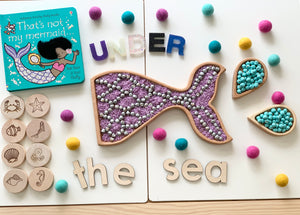 Mermaid tail sensory tray filled with purple beads and silver balls, wooden sea animal tokens, and colorful felt balls on white table