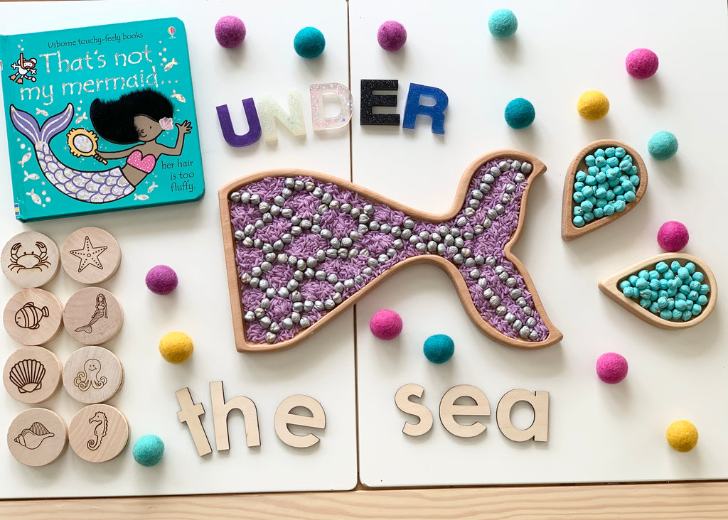 Mermaid tail sensory tray filled with purple beads and silver balls, wooden sea animal tokens, and colorful felt balls on white table