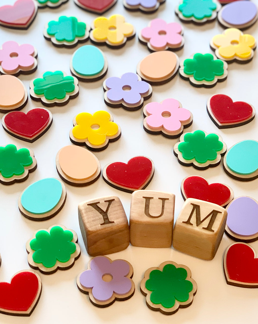 Wooden cookie-shaped pieces in flowers, hearts, eggs, and clovers with wooden letter blocks spelling YUM on white background
