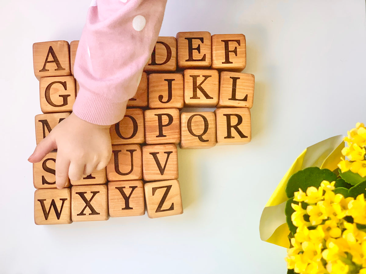 Natural 1.5-inch wooden alphabet blocks with engraved capital letters arranged with child’s hand and yellow flowers on white surface