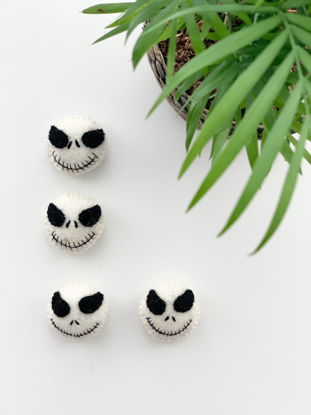 Four small handmade felt white skulls with black facial features arranged near green potted plant on white background