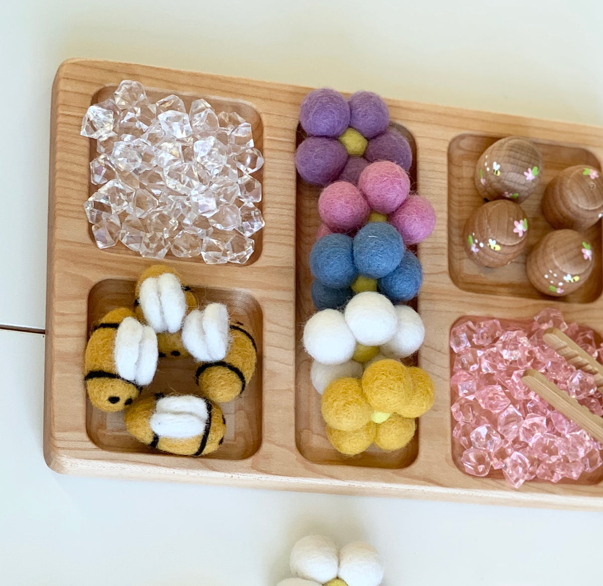 Natural wooden rectangle tray with compartments holding clear beads, felt flowers, felt bees, pink beads, and wooden cones