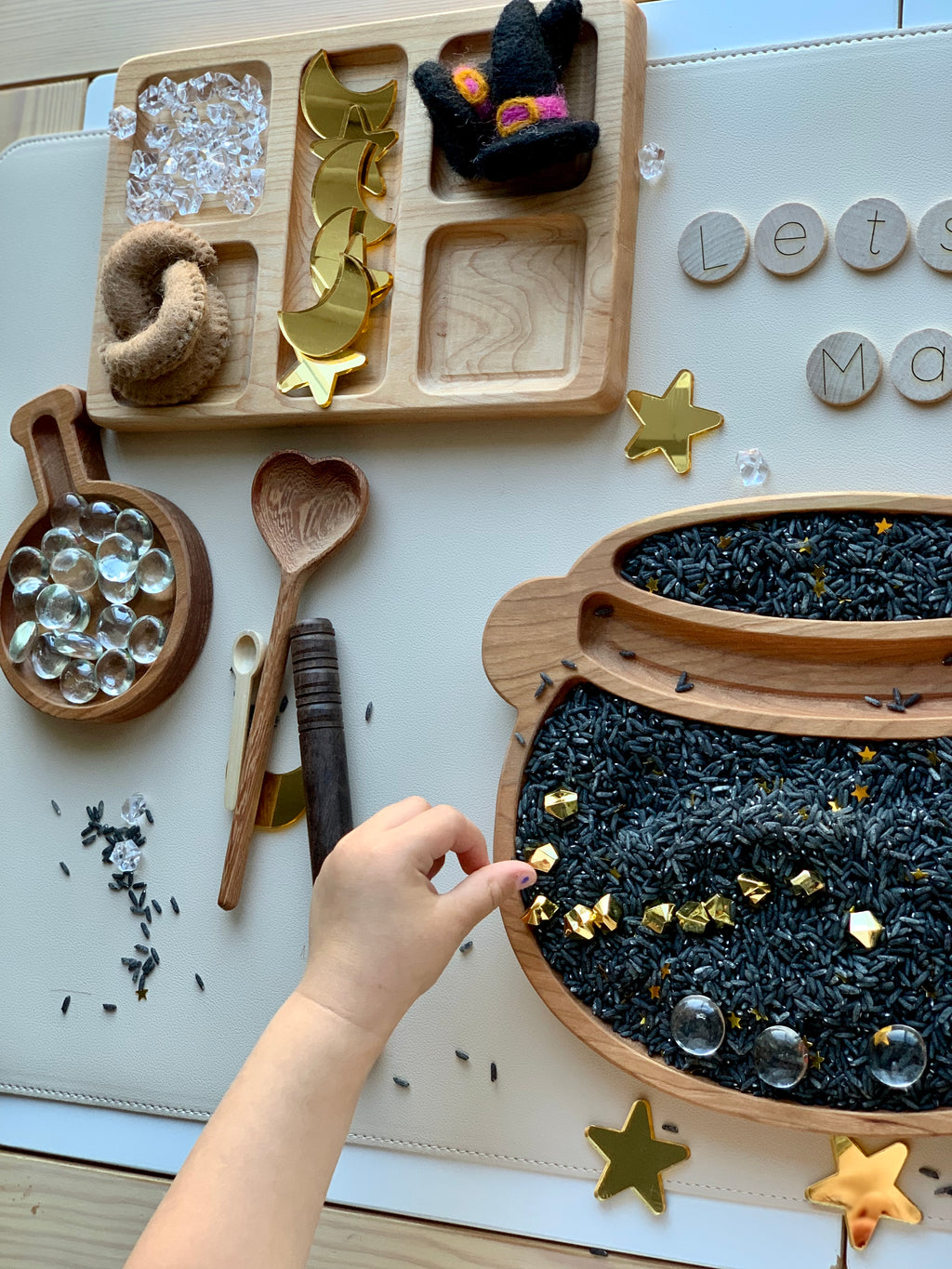 Small child’s hand playing with black dyed rice sensory bin filler with gold star confetti and wooden scoops on table