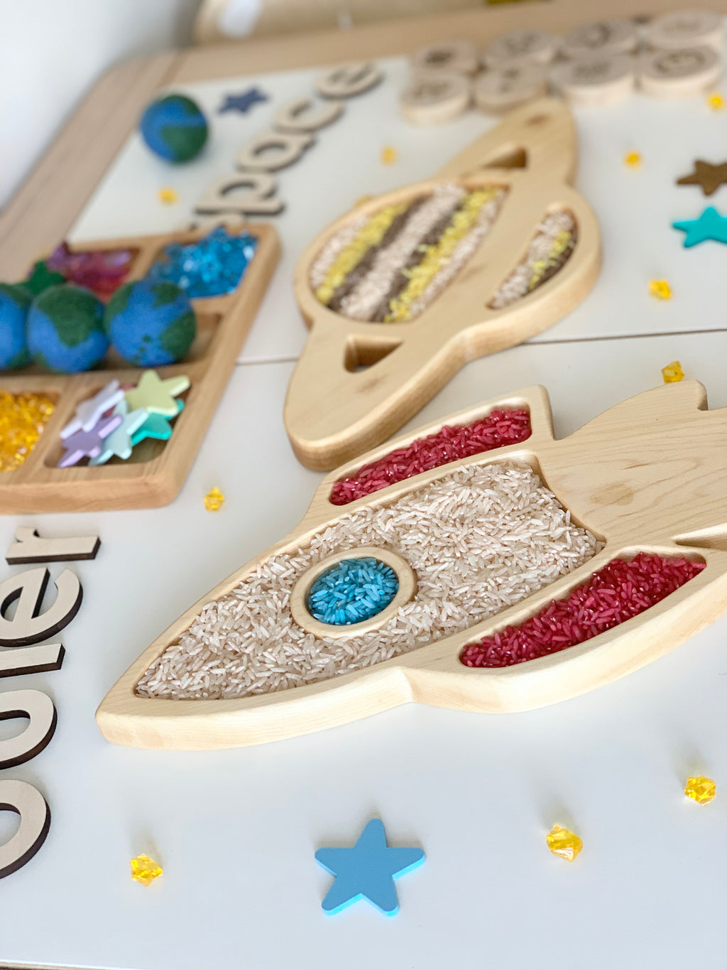 Wooden rocket ship sensory tray filled with colorful rice on white surface with star-shaped pieces