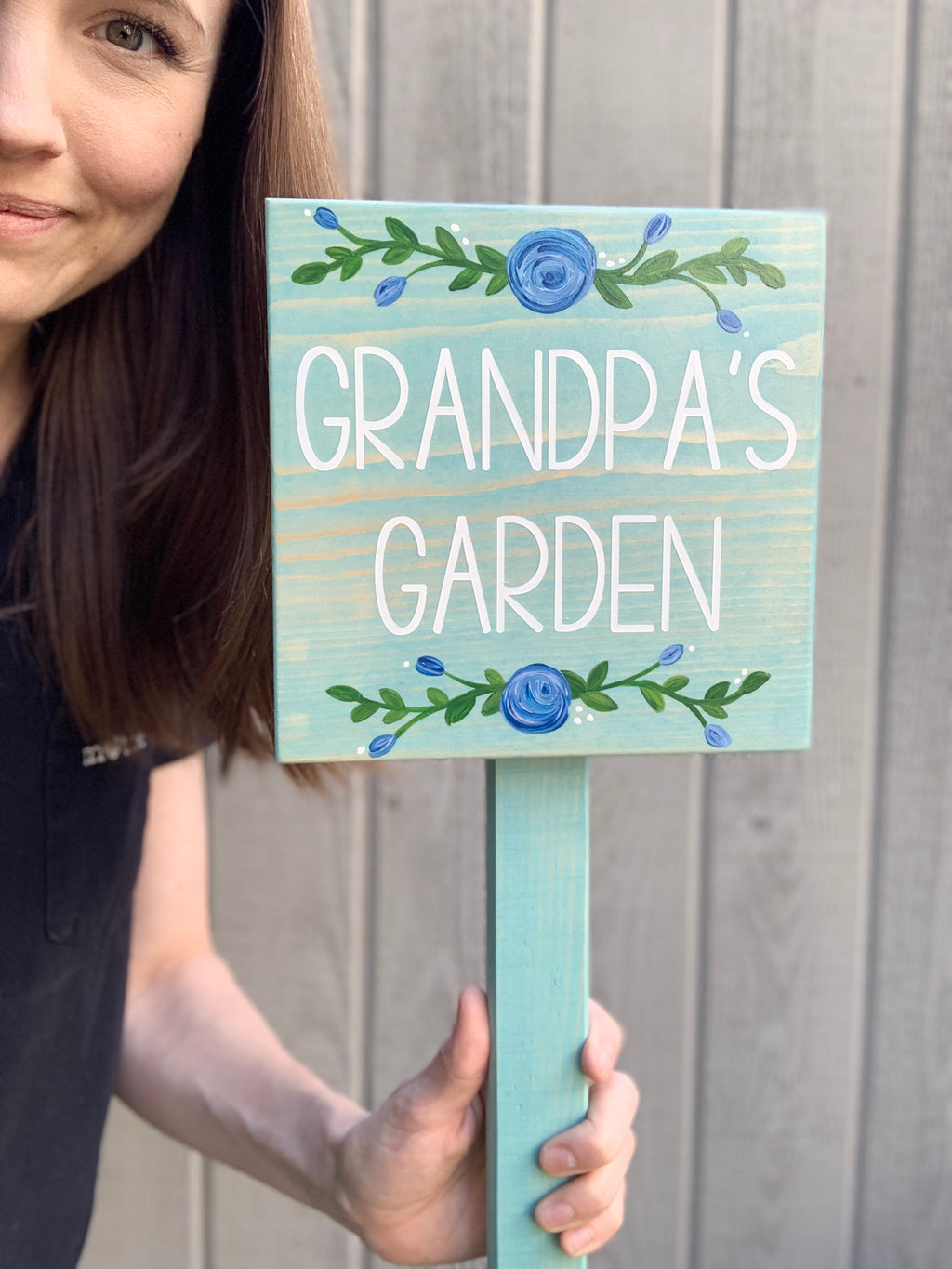 Small square wooden garden sign painted turquoise with white letters and blue floral accents held by woman