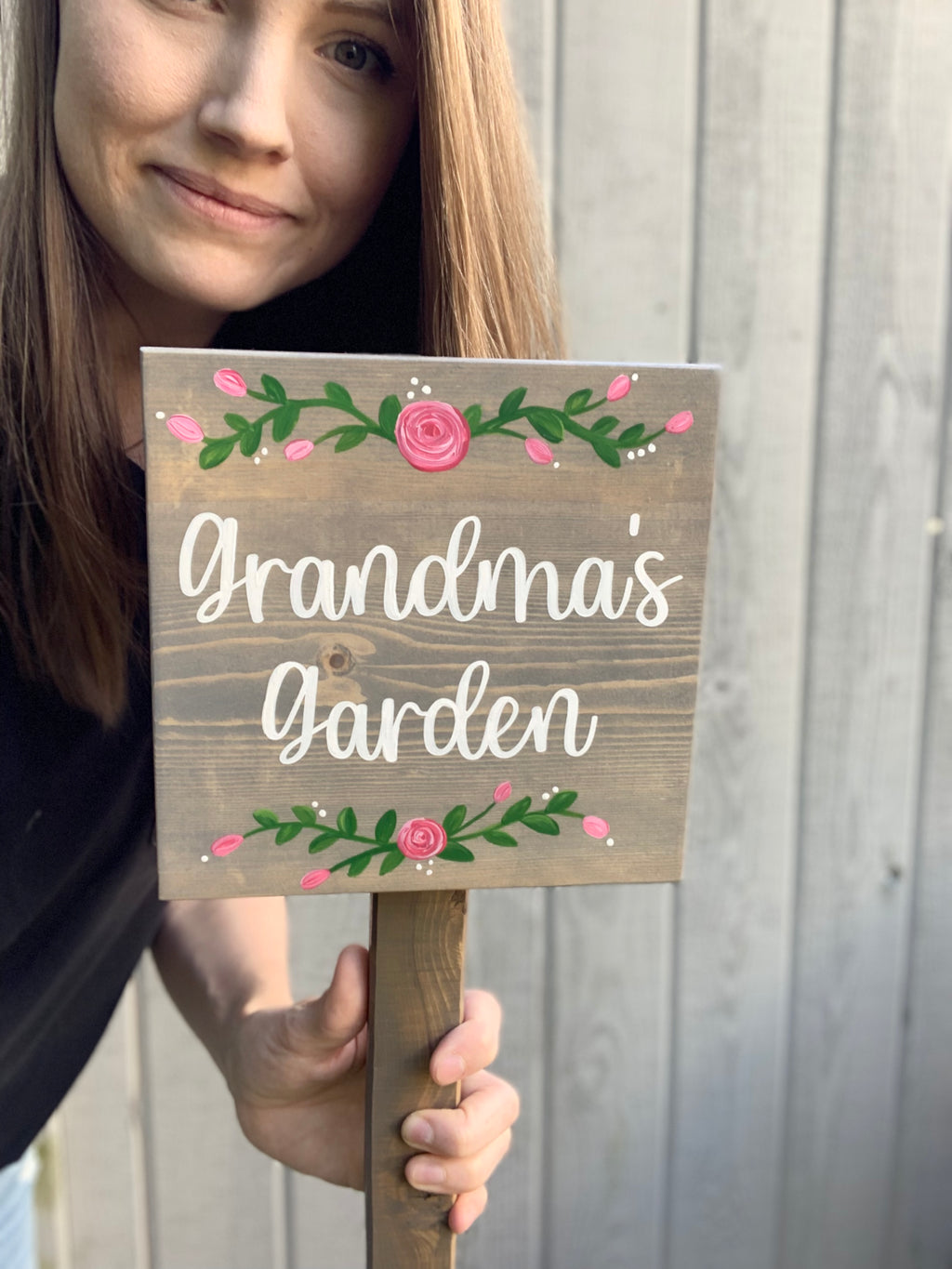 Small square wooden garden sign painted with white cursive letters and pink floral accents on natural wood background