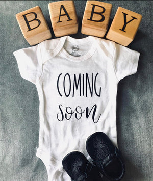 Natural wooden 1.5 inch blocks spelling BABY with white infant onesie reading coming soon and black baby moccasins on gray surface
