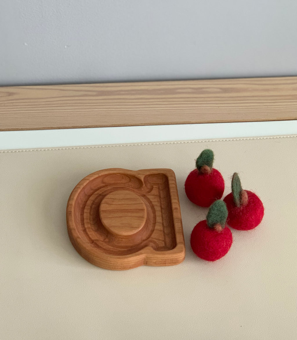 Natural wooden alphabet plate carved with lowercase letter a beside three red felt apples on cream surface