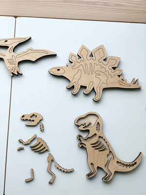 Natural wooden dinosaur skeleton puzzle pieces including T-Rex, Stegosaurus, and Pterodactyl on white table surface