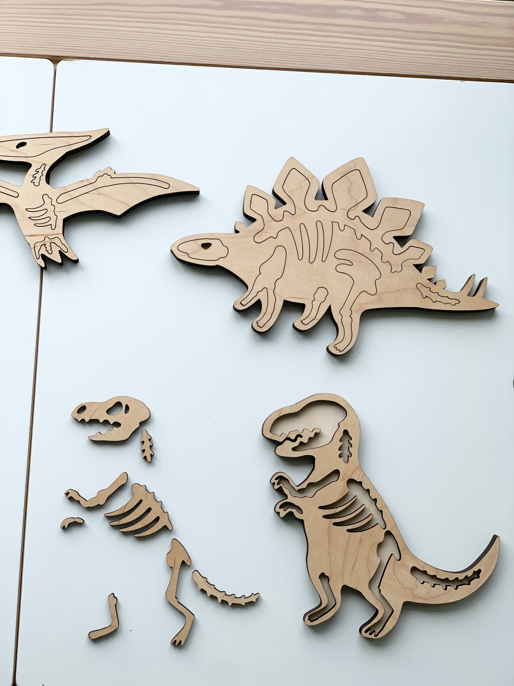 Natural wooden dinosaur skeleton puzzle pieces including T-Rex, Stegosaurus, and Pterodactyl on white table surface