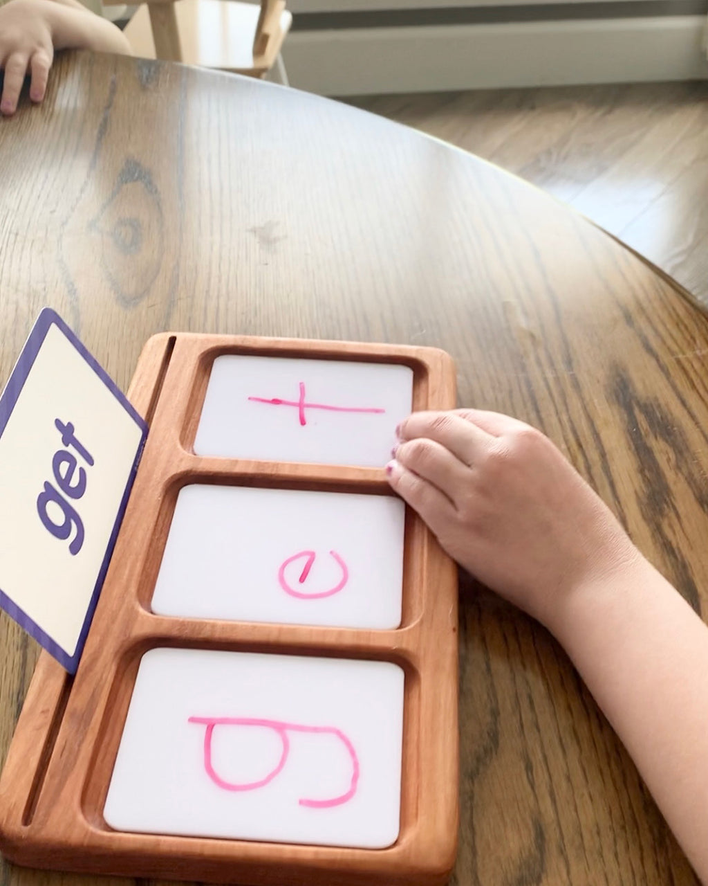 Child's hand writing letters g e t on three section wooden dry erase sensory tray on wooden table