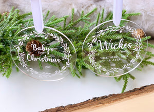 Two clear 1/8 inch acrylic Christmas ornaments with white engraved floral wreaths and personalized text hanging on white ribbons