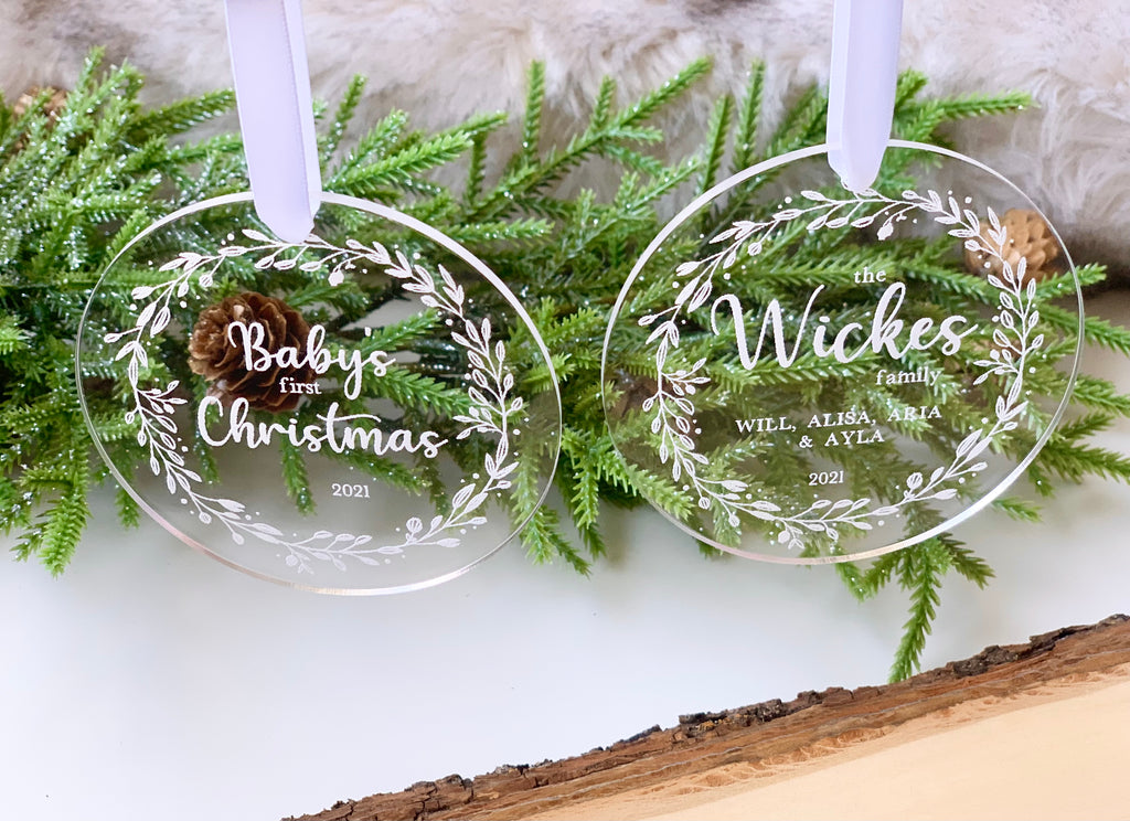 Two clear 1/8 inch acrylic Christmas ornaments with white engraved floral wreaths and personalized text hanging on white ribbons