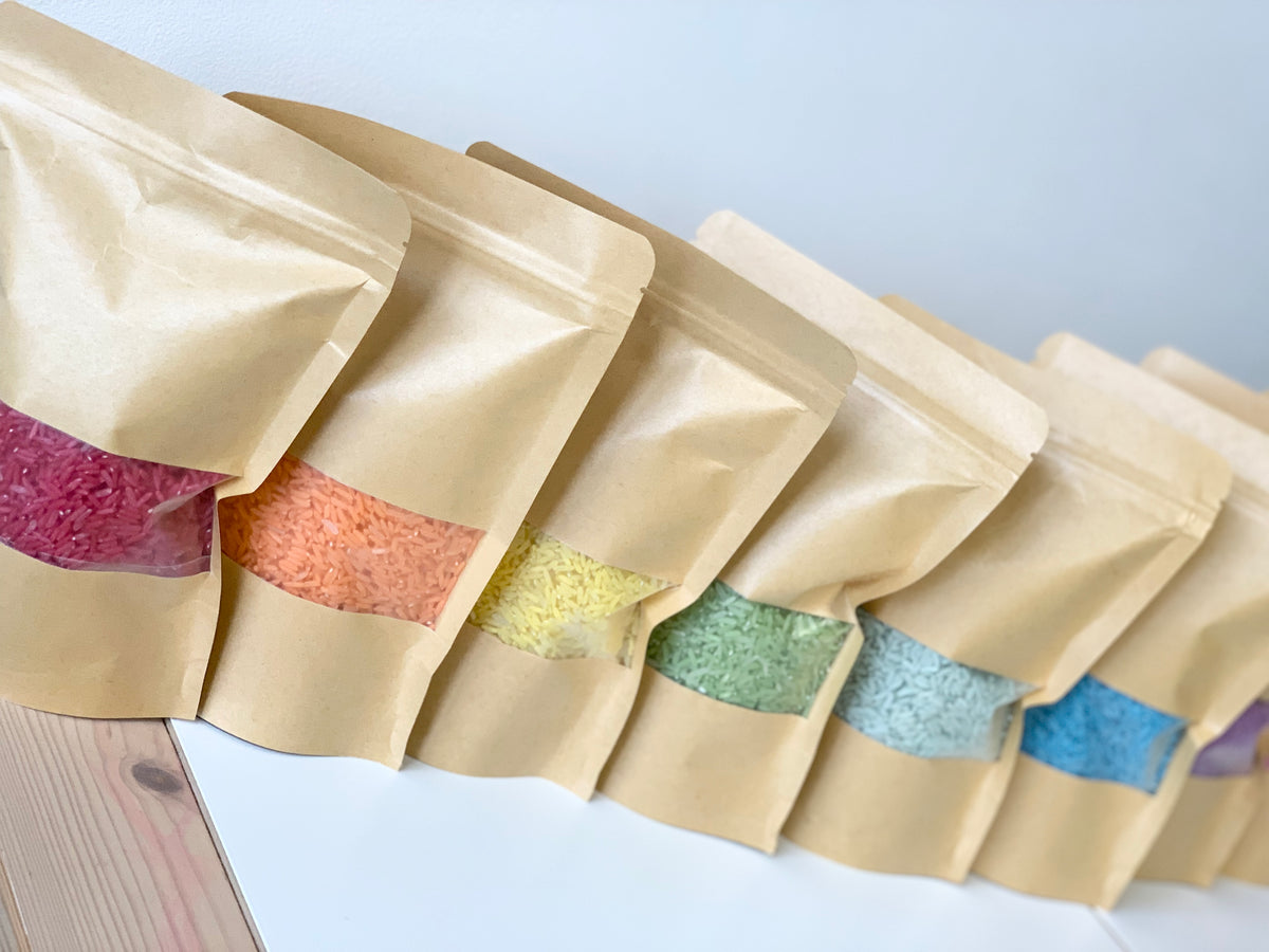 Seven kraft paper bags filled with different coloured sensory filler rice arranged in a row on white surface
