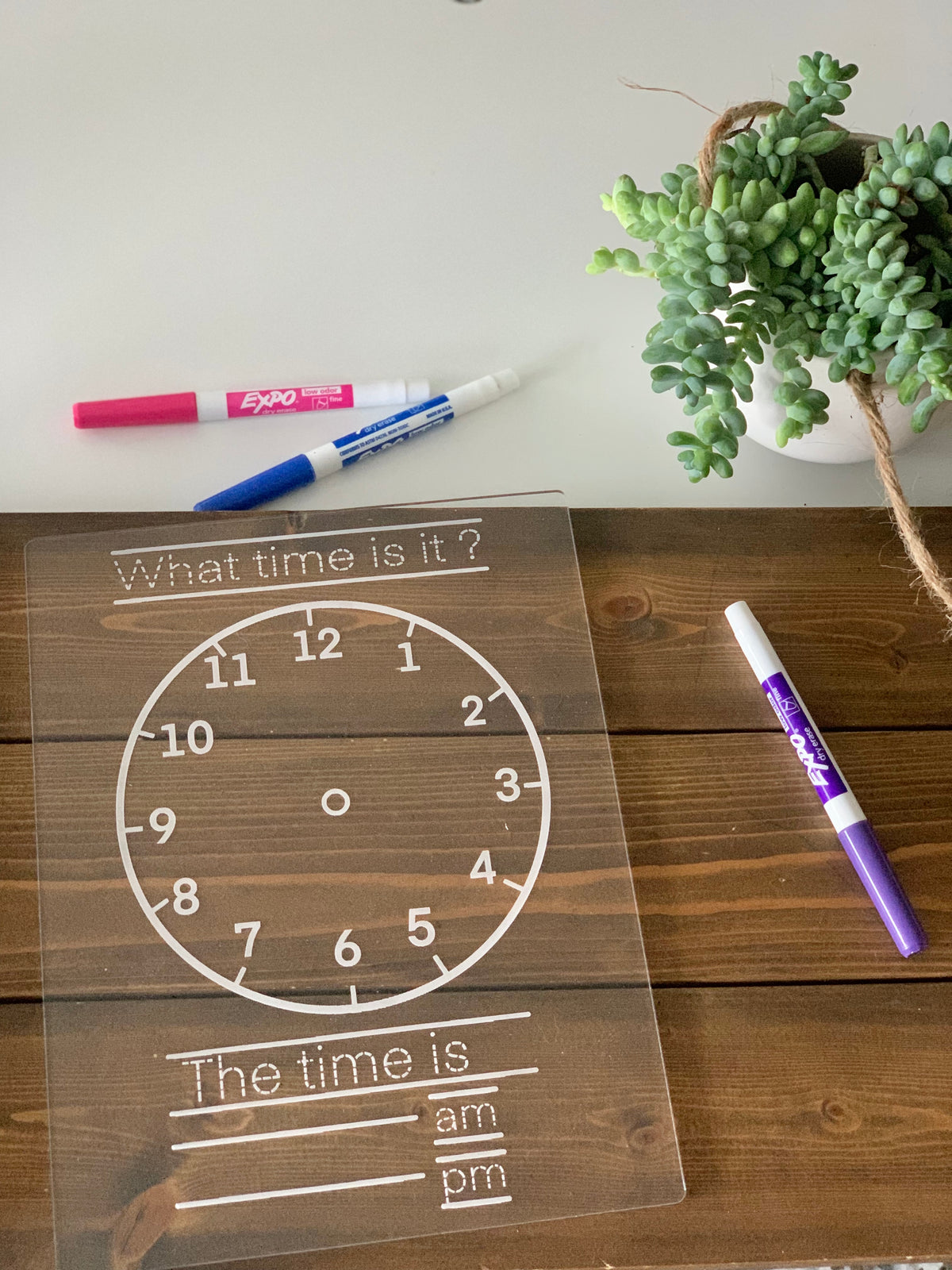 Clear acrylic dry erase board with analog clock and time writing practice sections on wooden surface with markers and plant