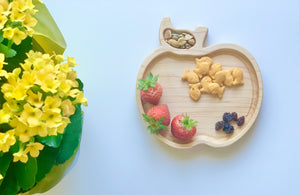 Natural wooden apple-shaped sensory tray with strawberries, goldfish crackers, raisins, and mixed nuts on white surface