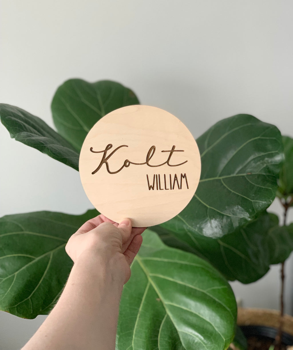 Hand holding a round light wood baby name announcement disc with name Kolt William in cursive and block letters