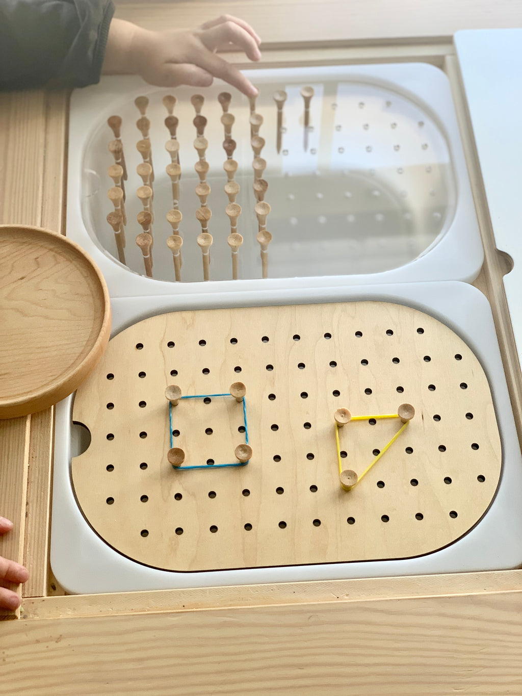 Small wooden pegboard with elastic bands forming shapes and wooden pegs arranged in clear storage tray on wood surface