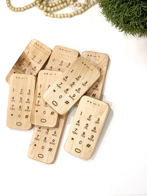Set of engraved wooden toy phones with numeric keypad details on light wood grain background near green plant and beads