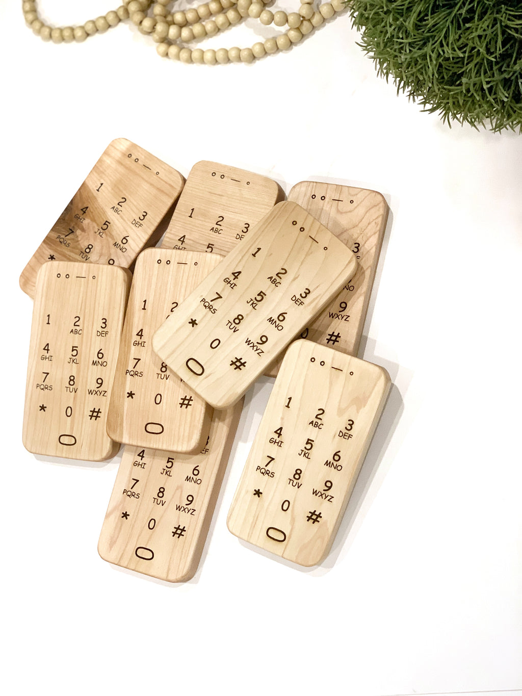 Set of engraved wooden toy phones with numeric keypad details on light wood grain background near green plant and beads