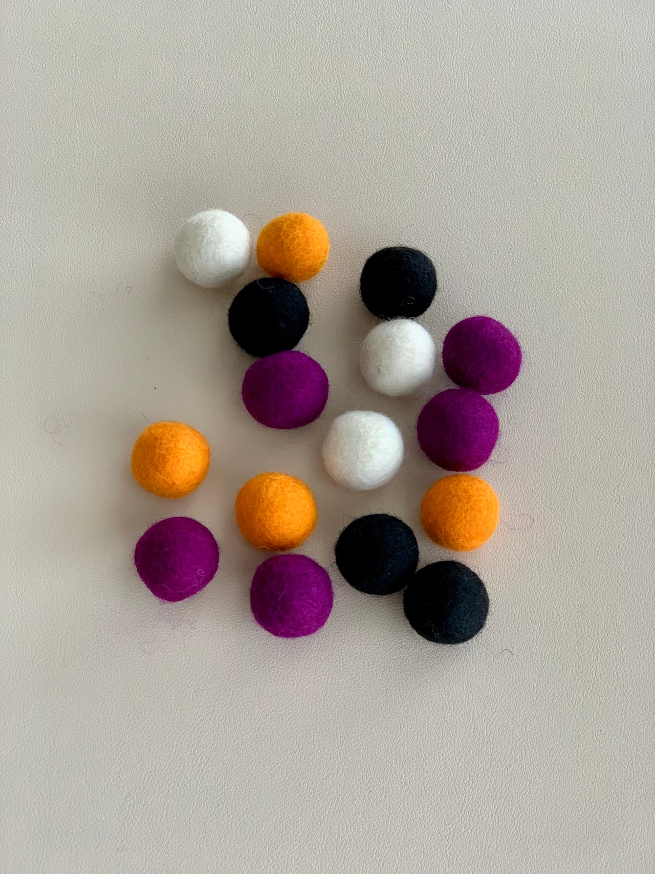 Sixteen small black, orange, purple, and white felt balls arranged on a light surface for Halloween craft use