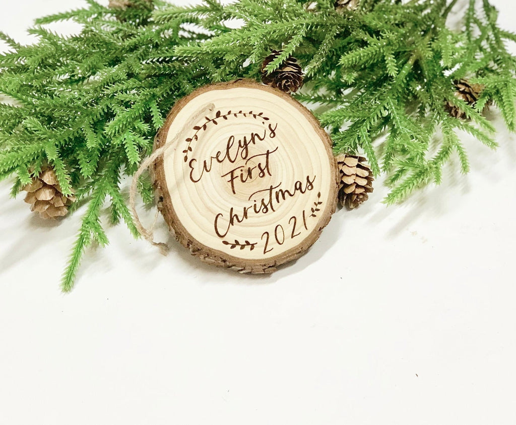 Wood burned wooden ornament engraved with Evelyn’s First Christmas 2021 surrounded by pine branches and pinecones on white background
