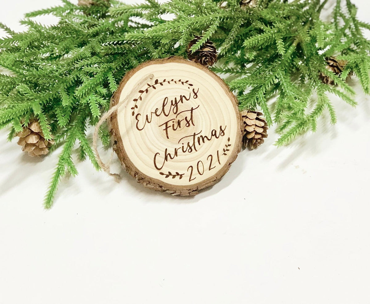 Wood burned wooden ornament engraved with Evelyn’s First Christmas 2021 surrounded by pine branches and pinecones on white background