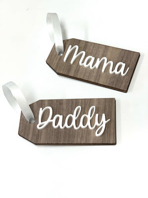 Two custom wooden gift tags with white cursive "Mama" and "Daddy" lettering and white satin ribbons on white background
