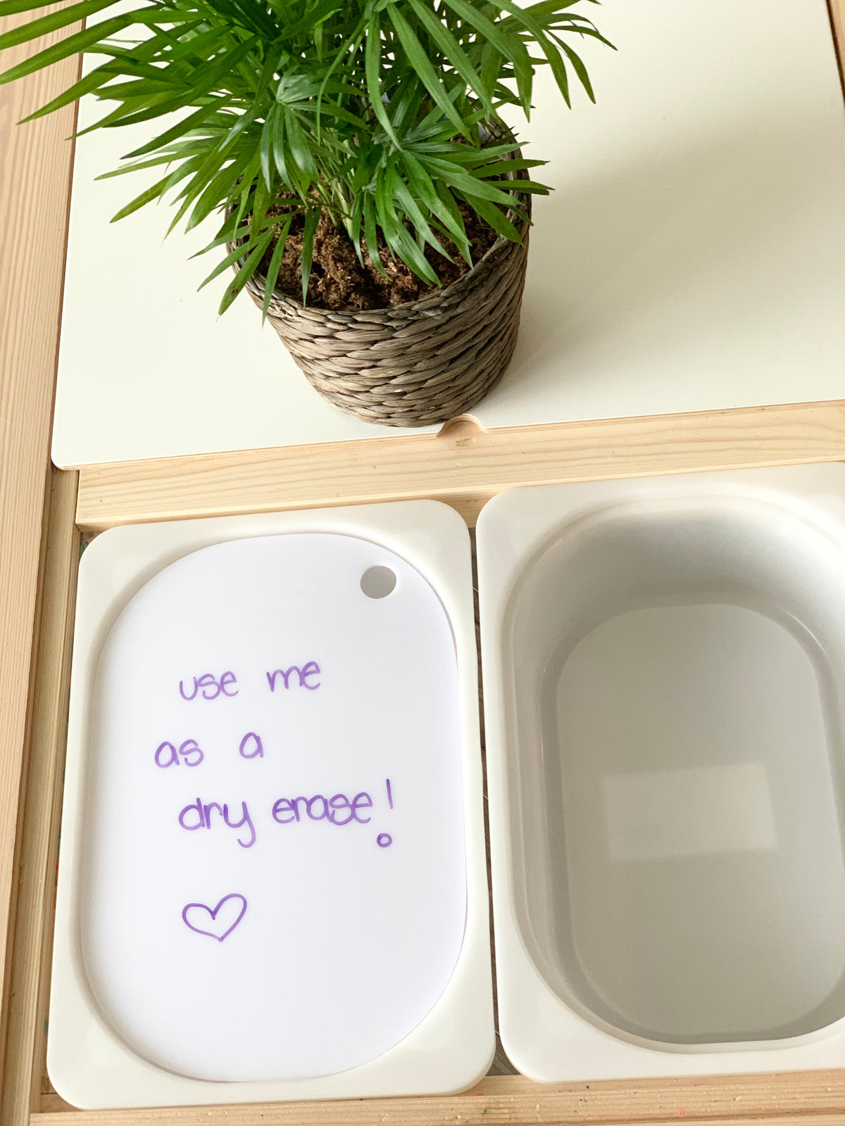 White acrylic Flisat table bin lid with purple dry erase marker writing and small green potted plant on light wood table