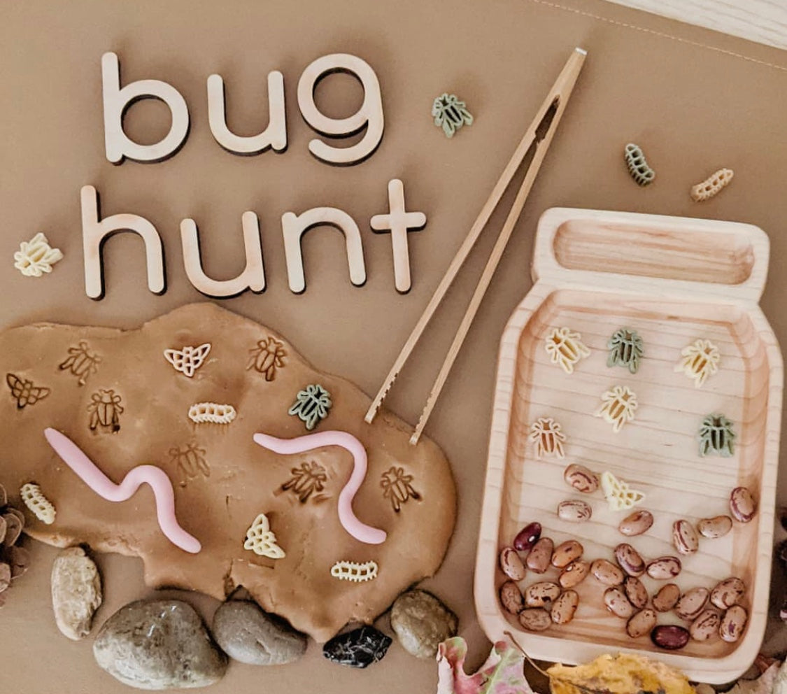 Small wooden jar-shaped sensory tray with tweezers, pasta bugs, beans, rocks, and "bug hunt" wooden letters on brown surface