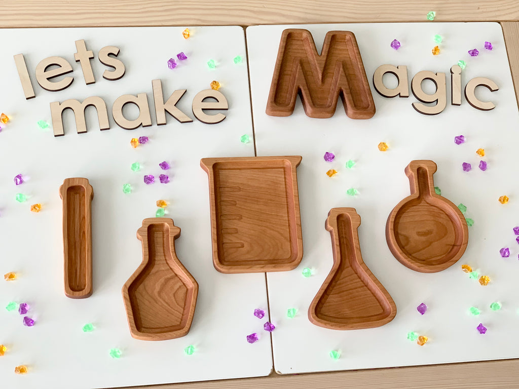 Wooden sensory trays shaped like science beakers and test tubes arranged on white boards with wooden letters spelling "lets make Magic
