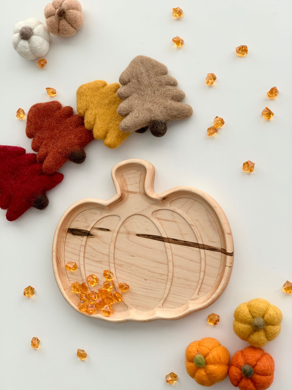 Light natural wooden pumpkin-shaped plate with engraved details surrounded by felt autumn leaves and pumpkins on white surface
