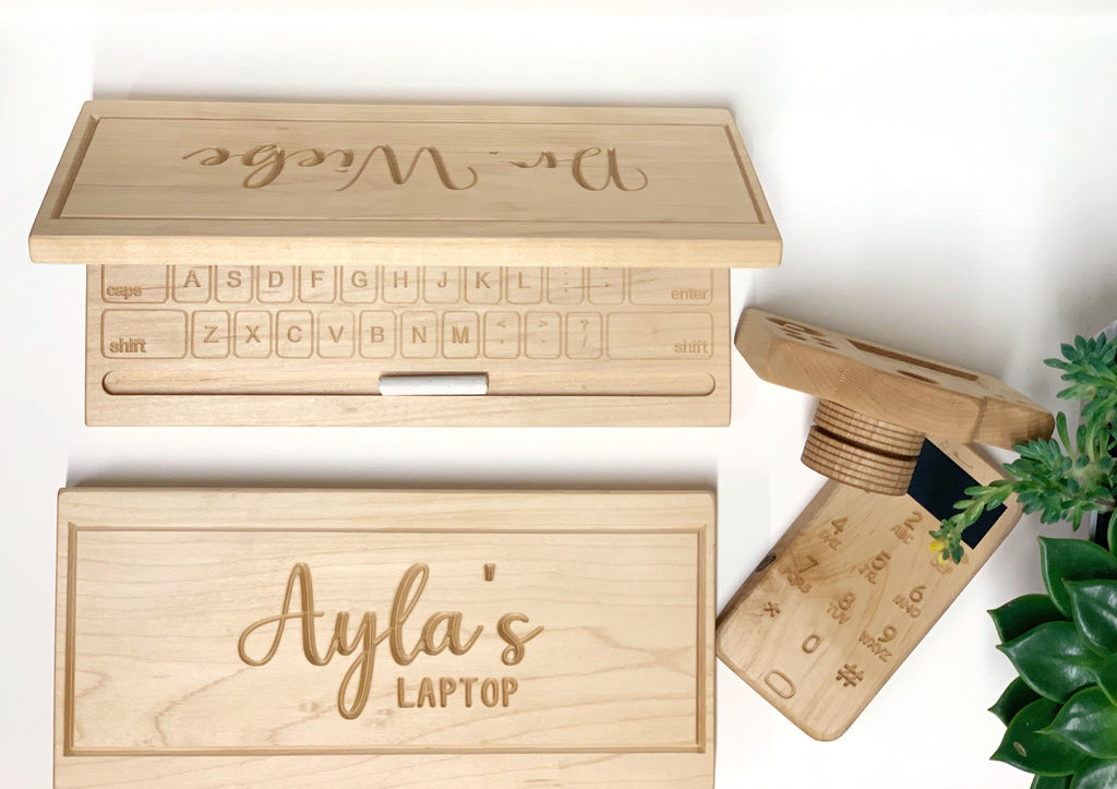 Natural wooden toy laptop with engraved keyboard and personalized lid alongside a rotating lens wooden toy camera on white surface