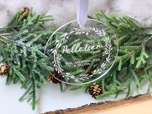Clear acrylic Christmas ornament engraved with family name and decorative leaf wreath hanging with ribbon over pine branches