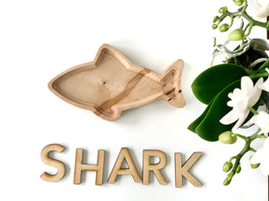 Natural wooden shark-shaped sensory tray with smooth edges displayed next to wooden letters spelling SHARK on white surface