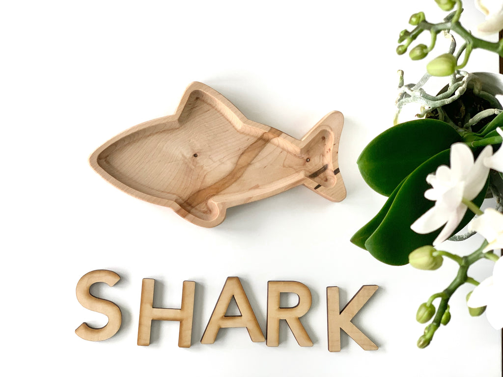 Natural wooden shark-shaped sensory tray with smooth edges displayed next to wooden letters spelling SHARK on white surface
