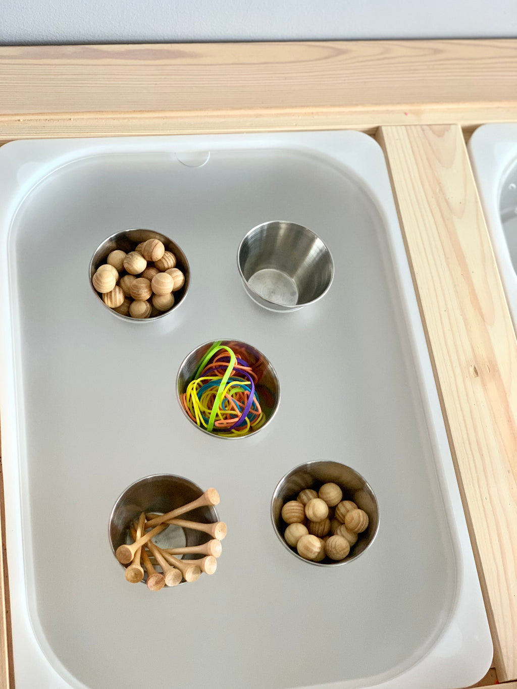 White acrylic insert with four metal cups holding wooden balls, pegs, and colorful rubber bands on light wood frame