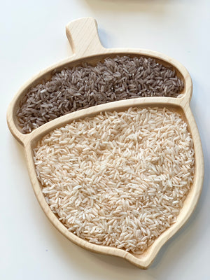 Natural wooden acorn-shaped sensory tray divided into two sections filled with light and dark rice grains on white surface