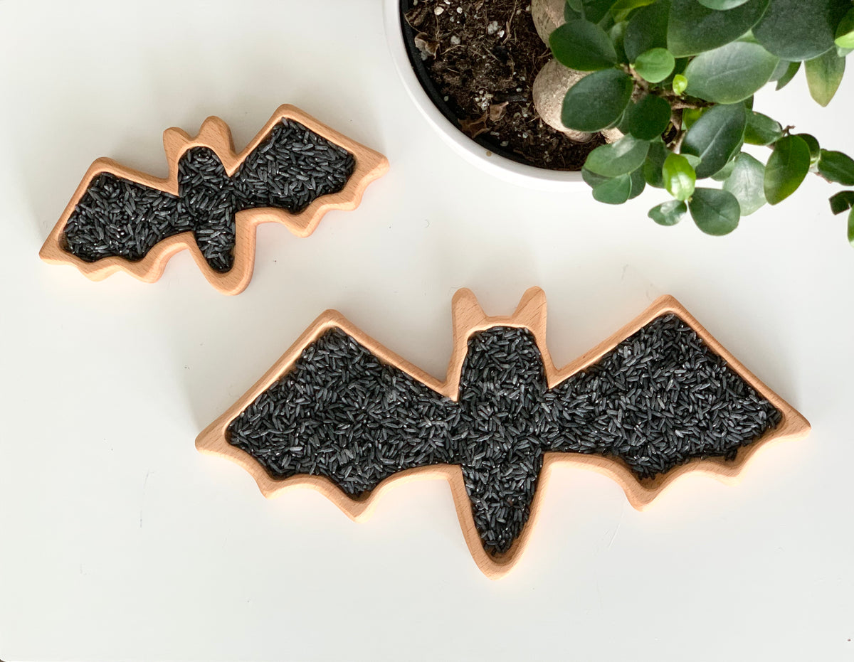 Two wooden bat-shaped sensory trays filled with black rice on white surface near green potted plant