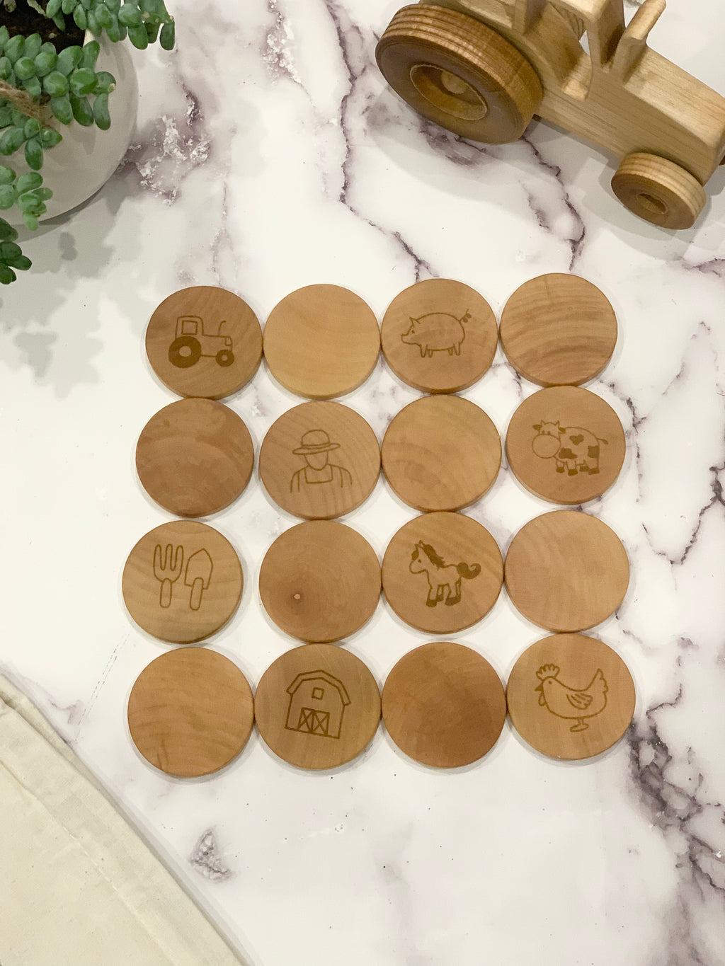 Round wooden memory game pieces engraved with farm icons arranged on white marble surface next to wooden tractor toy