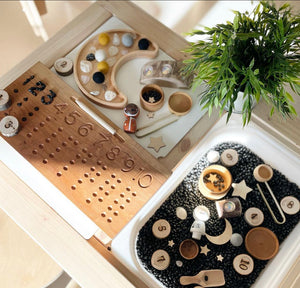 Wooden sensory tray with numbered pegs and moon-shaped plate holding small toys and black beads near green plant