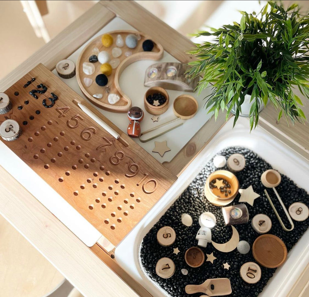 Wooden sensory tray with numbered pegs and moon-shaped plate holding small toys and black beads near green plant