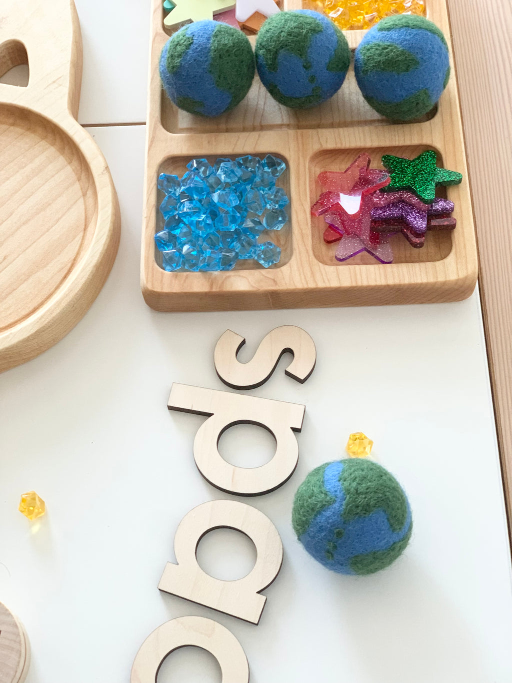 Four small handmade felt globes with blue and green continents on white surface near wooden letters and colorful craft gems