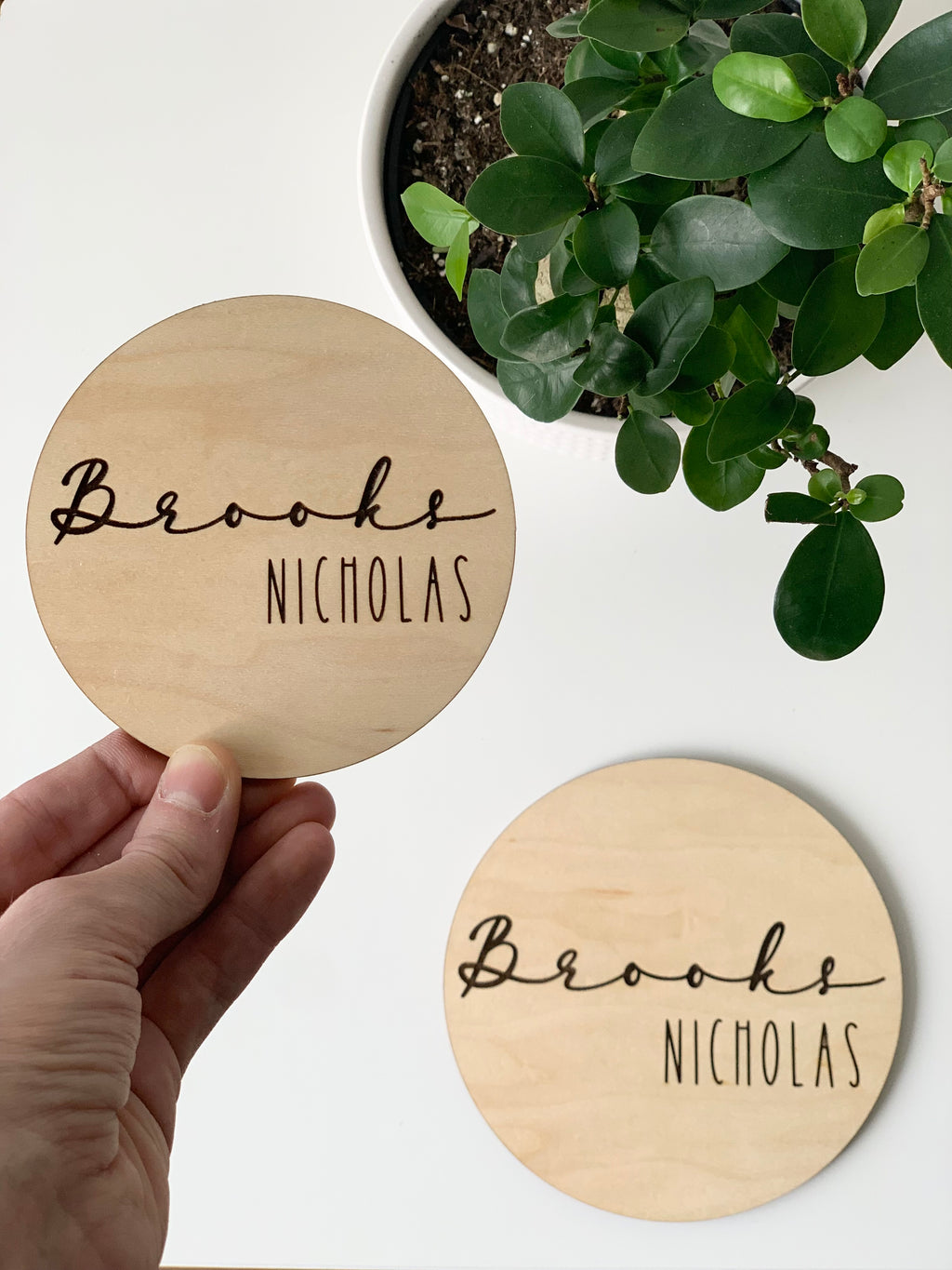 Round baby name announcement disc with cursive first name and capitalized middle name on natural maple wood background