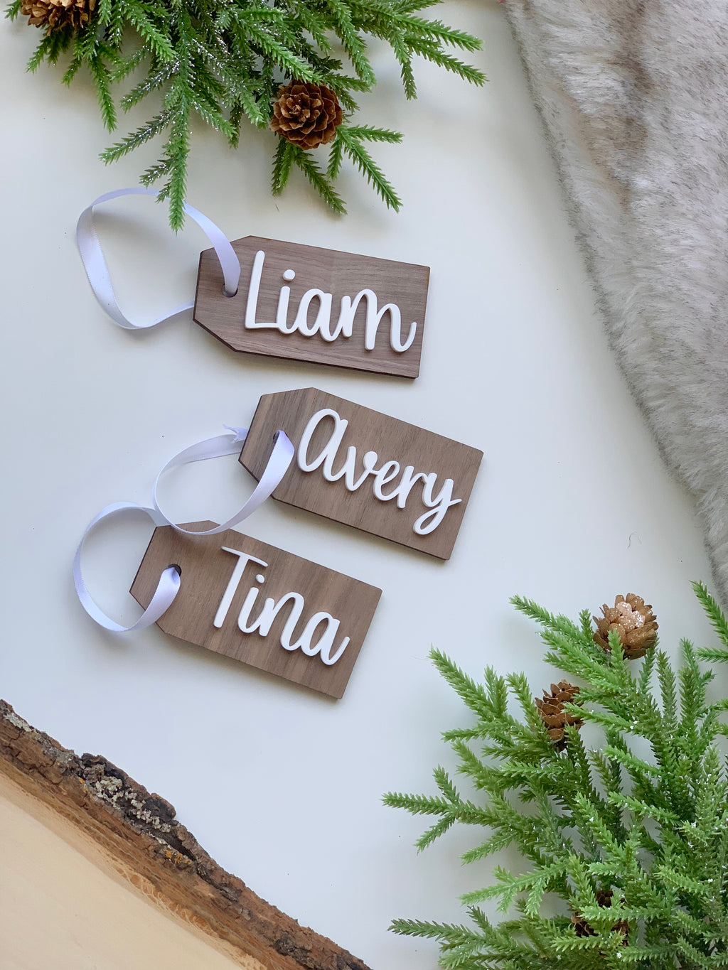 Three custom wooden Christmas gift tags with white cursive names Liam Avery and Tina on white surface with greenery and fur accents