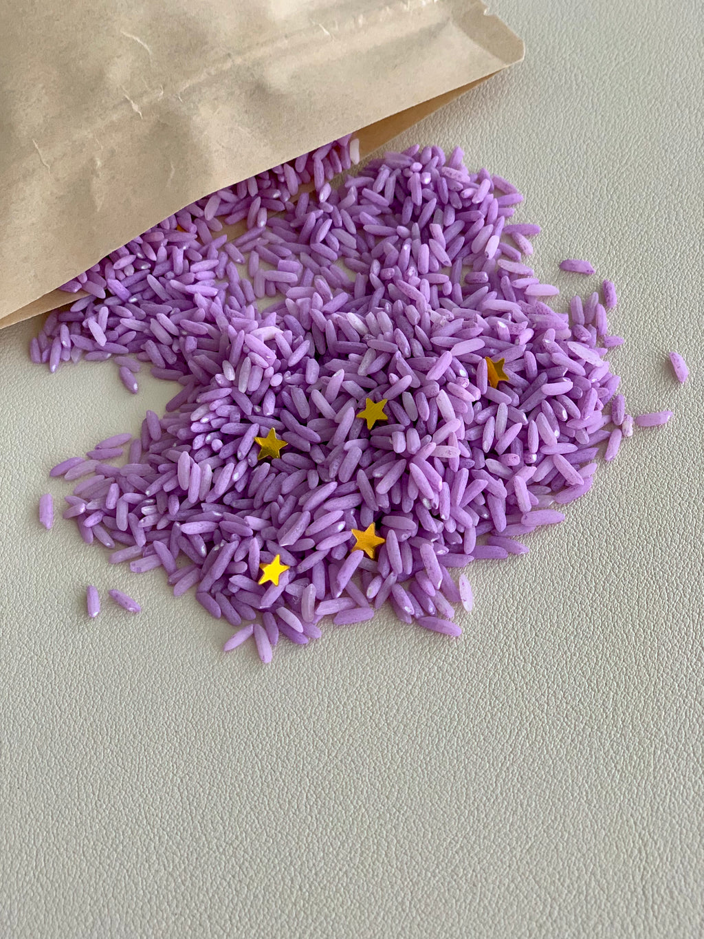 Small pile of dyed purple rice with gold star confetti spilling from brown paper bag on light surface